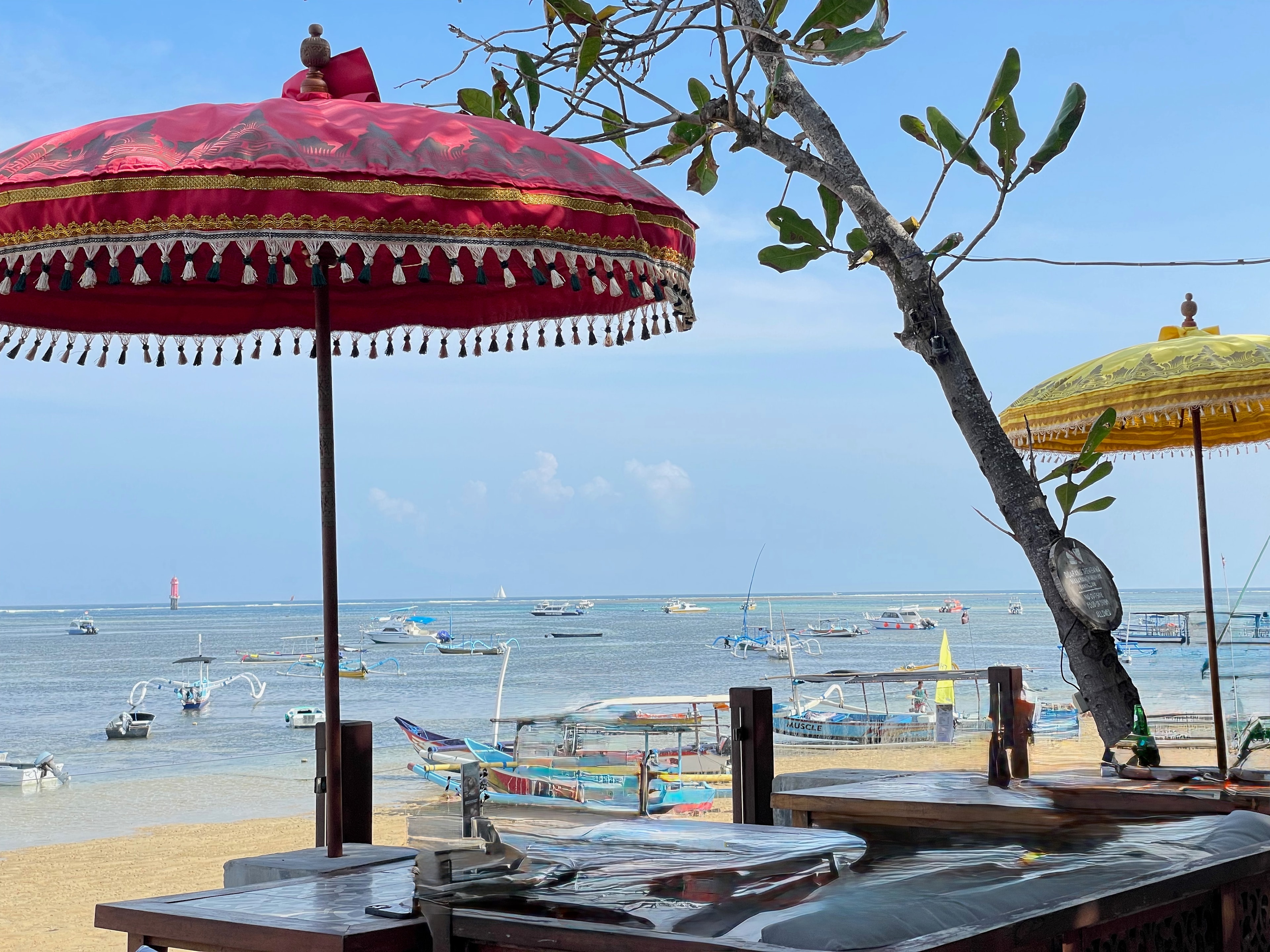 Balinese parasols over a beach table, boats in the bay