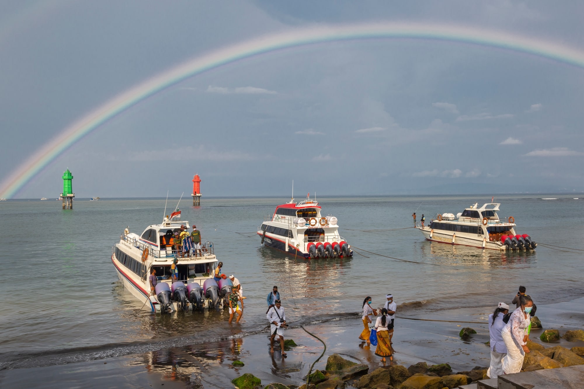 Three fast boats moored off the beach at Sanur under a rainbow, people in white ceremonial dress at the water's edge