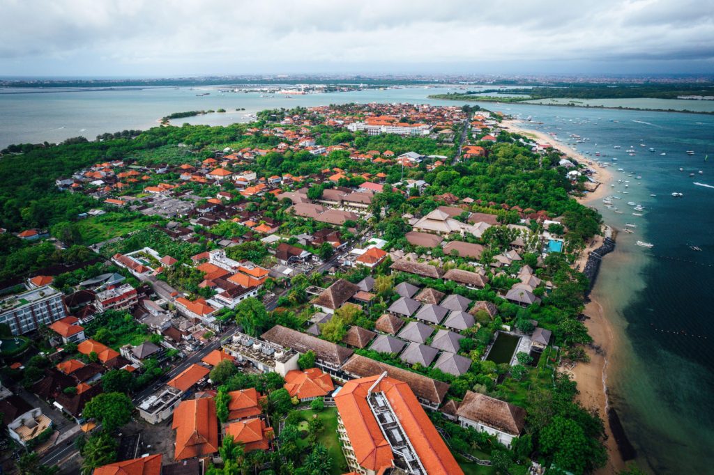 Aerial view of coastal town in Bali with traditional red-roofed buildings and lush greenery.